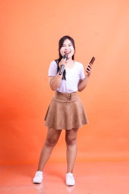 Elinde akıllı bir telefonla mikrofonla şarkı söyleyen gülümseyen genç bir kadın. Tam vücut çekim, turuncu arka planda izole.