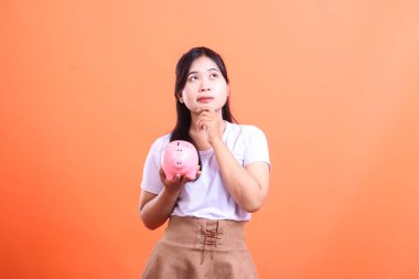 Beyaz tişörtlü genç düşünceli Asyalı kadın elinde kumbarayla finans durumunu ve turuncu arka planda izole edilmiş gelecek yatırımlarını düşünüyor..