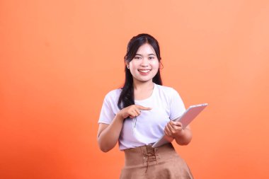 Elinde tabletle ekrandaki bir şeyi gösteren ve kameraya gülümseyen genç bir kadın. turuncu arkaplanda izole