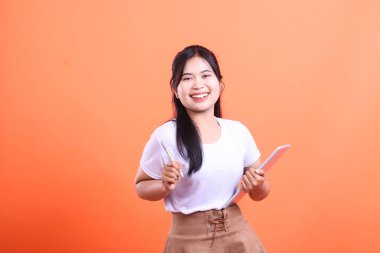 Tablet ve kalem tutan neşeli Asyalı genç bir öğrenci. Parlak bir şekilde gülümsüyor ve turuncu arka planda izole edilmiş bir şekilde doğrudan kameraya bakıyor..