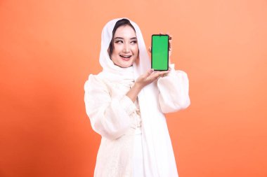 Mutlu bir Müslüman kadın tesettürlü akıllı telefonu yeşil ekranlı sunar. Uygulamaları ve mobil teknolojiyi sergilemek için mükemmeldir. turuncu bir arkaplanda izole.