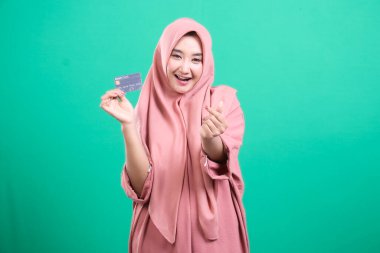 Tesettürlü, tesettürlü, tesettürlü genç Müslüman bir kadın yeşil arka planda izole bir şekilde parmaklarıyla kalp hareketi yaparken mutlu bir şekilde kredi kartını gösteriyor. Satın alma konusunda neşelidir..