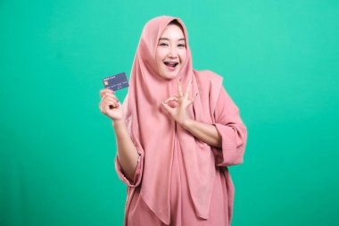 Elinde kredi kartı olan tesettürlü genç müslüman bir kadın yeşil arka planda izole edilmiş finansal hizmetlerden memnuniyetini dile getiriyor..