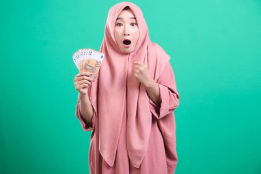 Heyecanlı, tesettürlü Müslüman bir kadın, yeşil arka planda izole edilmiş mali kazancı için şaşırdığını ve heyecanını dile getiren avro banknotlarına hayran..