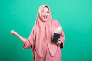 Heyecanlı tesettürlü Müslüman kadın, yeşil bir arka planda izole edilmiş, cüzdanında avrolar tutan ve yumruğunu heyecanla pompalayan mali başarısını kutluyor. Bu refah ve serveti resmediyor..