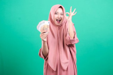 Elinde parası olan mutlu Müslüman kadın yeşil bir zeminde izole bir şekilde mali başarısını ve zenginliğini kutluyor. Elindeki kazançlarla başarı ve neşe saçar..