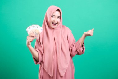 Avro banknotları tutan tesettürlü gülümseyen Müslüman kadın, yeşil bir arka planda izole edilmiş ekonomik özgürlük ve yatırım umutları sergiliyor. Resim başarılı mali güvenlik ve zenginlik refahını vurguluyor.