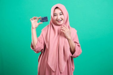 Gülümseyen bir kredi kartı tutan mutlu Müslüman kadın, yeşil arka planda soyutlanmış modern bankacılık erişimini gösteriyor. Finansal anlaşmalara ve dijital ticarete olan güvenini gösteriyor..