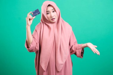 Kafası karışmış tesettürlü bir kadın omuz silkerken bir banka kartı tutuyor ve yeşil arka planda izole edilmiş mali durumdaki kafa karışıklığını dile getiriyor. Belirsiz görünüyor, belki online alışveriş sorunları ya da kredi sorunları yüzünden..