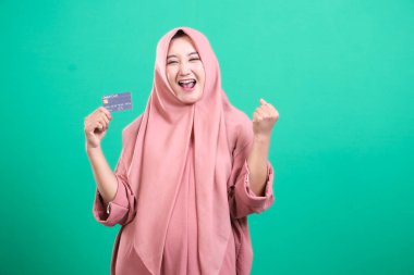 Tesettürlü neşeli kadın bir kart tutuyor, başarılı bir alışverişi kutluyor ve yeşil bir arka planda izole edilmiş, kazanan bir jestle mutluluğunu ifade ediyor. Kullanım kolaylığı ve dijital ilerlemenin gösterimi.