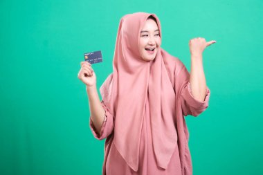 Tesettürlü gülümseyen Müslüman bir kadın umut verici bir şeye işaret ederken sevinçle kredi kartı sunar. Yeşil arka planda izole edilmiş bu, finansal fırsatları ve modern yaşamı temsil ediyor..