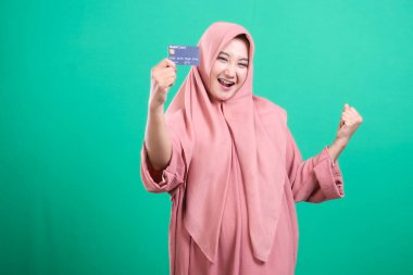 Bir tesettürdeki neşeli Müslüman kadın finansal başarısını bankamatik kartıyla kutluyor, saf hazzı ifade ediyor, yeşil bir arka planda izole edilmiş, modern bankacılık sergiliyor..