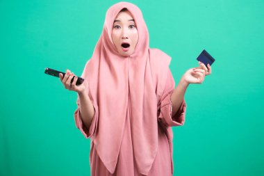Başörtüsü içinde genç bir Müslüman kadın, elinde telefon ve kredi kartı tutarak, yeşil arka planda izole edilmiş modern finansmanı ima ederek, kültür ve teknolojinin entegrasyonunu sembolize ederek şaşkınlığını dile getiriyor..