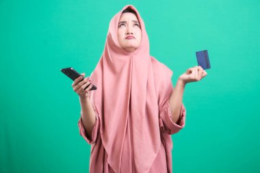 Elinde kredi kartı ve telefonla tesettürlü tedirgin müslüman bir kadın, mali konularda kafa karışıklığı gösteriyor, yeşil arka planda izole edilmiş. Resim, bir belirsizlik ve modern mali zorluklar hissi veriyor..