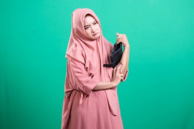 Başörtülü genç müslüman kadın endişeli bir ifadeyle boş cüzdanına bakıyor, yeşil arka planda izole edilmiş maddi sıkıntıları yansıtıyor..