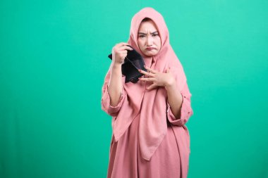 Mutsuz Müslüman kadın boş bir cüzdan gösteriyor. Finansal sorunlar ve yeşil arka planda izole edilmiş ekonomik belirsizlikten duyduğu hayal kırıklığını dile getiriyor..