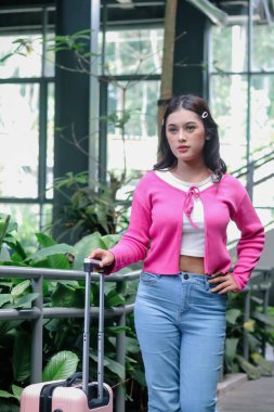 Parlak pembe hırka ve kot pantolon içinde maceraya hazır çekici genç bir kadın. Elinde pembe bir bavul tutuyor, doğal aydınlatılmış bir kalkış terminalinde şık bir ton çiziyor..