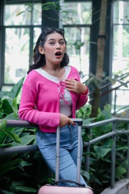 Genç bir kadın sürpriz bir yüz ifadesi sergiliyor. Günlük kıyafetler ve pembe hırka giyip, bavuluyla birlikte, kapalı, tropikal bir ortamda seyahat yolculuğuna çıkıyor..