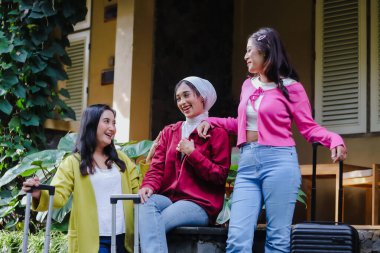 Üç mutlu genç kadın, her biri bavul taşıyan ve yolculuğa hazır neşeli bir şekilde bir araya gelmenin tadını çıkarıyor. Sahne doğal ışıkla yıkanır, canlı bir dostluk ve rahat bir an gösterir..