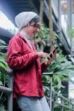 Başörtüsü takan genç bir kadın çantanın yanında cep telefonu kullanırken gülümsüyor, modern bir binada yeşil tropikal bitkiler yerleştiriliyor, dijital dünyayla kusursuz bir bağ kuruyor..