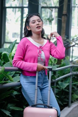 Canlı pembe bir hırka ve açık mavi kot giymiş dengeli genç bir gezgin, macerasında duraklıyor, sakin bir bahçe ortamında pastel bir bavul tutuyor. Seyahatinin ortasında bir an dinleniyor..