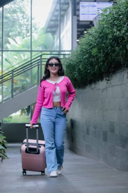 Neşeli genç bir kadın, parlak pembe bir hırka ve güneş gözlüğü giyiyor, tatlı tatlı gülümsüyor bavulunu yuvarlarken, yolculuğa ve dünyayı keşfetmeye hazır..