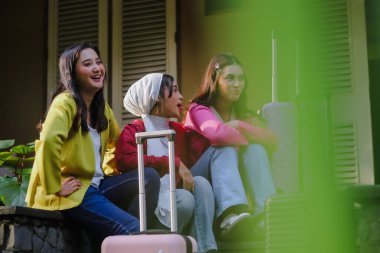 Üç şık genç bayan dışarıda bavullarıyla oturuyor, beklenti ve dostluk yayıyorlar. Gülüşlerini ve gülüşlerini paylaşıyorlar, birlikte tatile çıkmaya hazırlar..