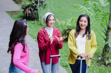 Bavullarını taşıyan üç mutlu genç bayan, açık havada canlı sohbete girişiyor. Üçlü tatildeyken gülüyorlar, birbirleri ile rahat görünüyorlar..