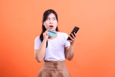 Elinde kredi kartıyla şaşırmış gibi görünen genç bir kadın ve kafası karışmış bir yüz ifadesi olan bir cihaz telefonu tutuyor. turuncu arkaplanda izole