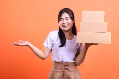 Elinde bir yığın karton kutu tutan gülümseyen bir kadın müşteriye teslimatı sunuyor, turuncu arka planda izole edilmiş..
