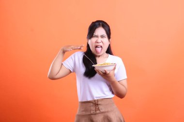 İğrenmiş bir ifadeyle genç bir kadın elinde bir kase erişteyle turuncu arka planda izole bir şekilde dilini dışarı çıkarıyor..