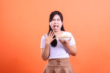 İğrenmiş bir ifadeyle genç bir Asyalı kadın, elinde bir kase erişteyle turuncu arka planda izole edilmiş bir şekilde,.