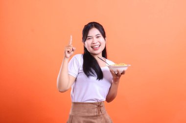 Neşeli Asyalı kadın elinde bir kase erişte tutarken gülümsüyor, parmağıyla turuncu arka planda izole edilmiş bir fikri işaret ediyor.