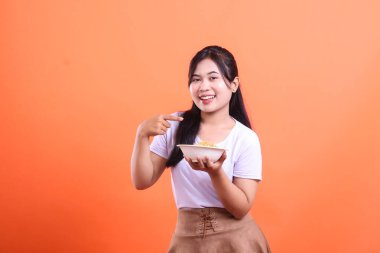 Büyük bir gülümsemesi olan genç bir kadın bir kase erişteyi mutluluk ve neşe ile sunuyor. turuncu arkaplanda izole