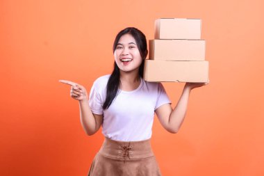 Karton kutular taşıyan neşeli genç bir kadın solu işaret ediyor, turuncu arka planda izole edilmiş..