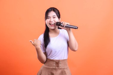 Genç, neşeli bir kadın mikrofona şarkı söylüyor, başparmağıyla işaret ediyor, mutlu ve hevesli görünüyor, turuncu arka planda izole edilmiş.