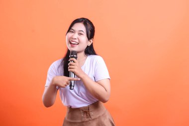 Elinde mikrofonla gülümseyen ve onu işaret eden bir kadın, turuncu arka planda izole edilmiş bir şov izlenimi veriyor.