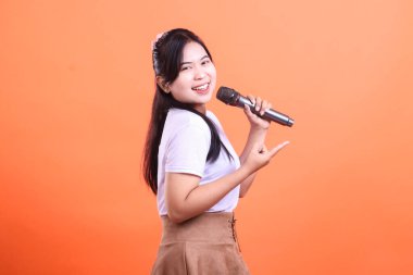 Güzel ve neşeli genç bir kadın mikrofon tutarken ve parmağıyla işaret ederken turuncu arka planda izole bir şekilde şarkı söylüyor.