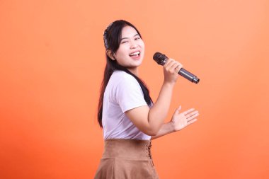Mikrofonlu genç bir Asyalı kadın sevinçle şarkı söylüyor, gülümsüyor ve sahnede elini gösteriyor, turuncu arka planda izole bir şekilde.