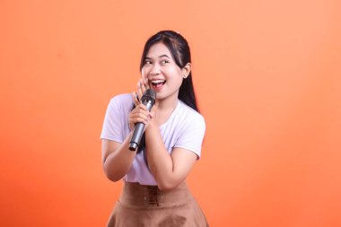 Gülümseyen genç bir kadın mikrofona sevinçle şarkı söylüyor, büyük bir mutluluk ve coşkuyla başka yere bakıyor, turuncu arka planda izole edilmiş.