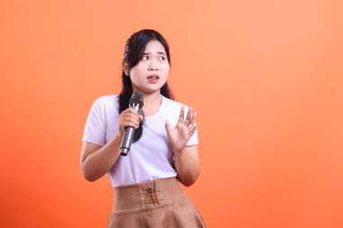 Genç bir Asyalı kadın, korkmuş ve şaşırmış görünüyor, mikrofon tutuyor, muhtemelen bir gösteri ya da röportaj için, turuncu arka planda izole edilmiş..