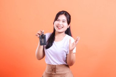 Mutlu genç bir kadın araba anahtarını büyük bir gülümsemeyle gösterir. Yeni araba sahipliğini kutluyor ve turuncu arka planda izole edilmiş neşeli bir ifadesi var.