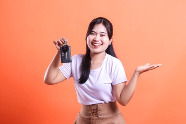 Araba anahtarı olan mutlu Asyalı kadın. Neşeli genç bir kadın araba anahtarını sunar ve turuncu arka planda izole edilmiş boş bir el alanı sunar.