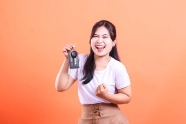 Genç, neşeli Asyalı bir kadın arabanın anahtarlarını elinde tutuyor, yeni bir araç ya da ev sahibi oluşunu mutlu bir ifadeyle kutluyor. turuncu arkaplanda izole