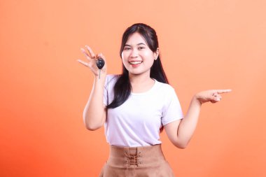 Gülümseyen Asyalı kadın arabanın anahtarlarını tutuyor ve sağ tarafı işaret ediyor, bir yön ya da terfi olduğunu gösteriyor. turuncu arkaplanda izole.