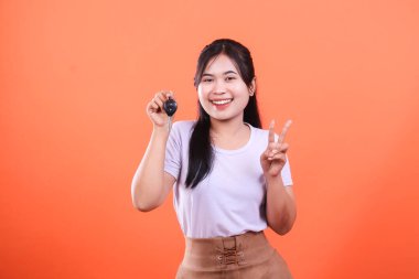 Neşeli bir Asyalı kadın arabanın anahtarlarını gösteriyor, gülümsüyor ve parmaklarıyla kameraya barış işareti yapıyor. turuncu arkaplanda izole.