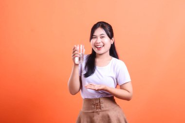 Elinde bir bardak su tutan gülümseyen Asyalı kadın susuzluğu ve sağlıklı yaşam tarzını teşvik ediyor. turuncu arkaplanda izole.