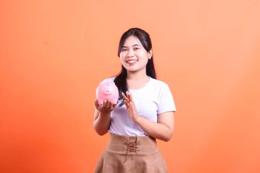 Elinde pembe bir kumbarayla gülümseyen genç bir kadın gelecek için tasarruf ve finansal planlamayı temsil ediyor. turuncu arkaplanda izole