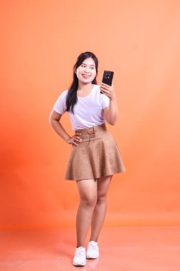 Güzel, genç bir kadın gülümsüyor, akıllı telefonuyla selfie çekiyor, tüm vücudunu çekiyor. turuncu arkaplanda izole.