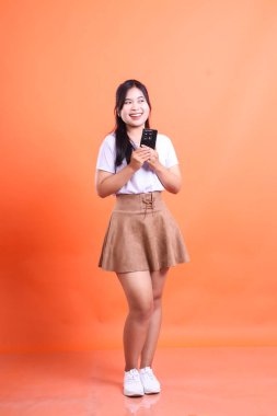 Akıllı telefon kullanan ve gülümseyen genç Asyalı güzel bir kadın turuncu arka planda izole edilmiş mutlu bir ifadeyle yüzünü çeviriyor.